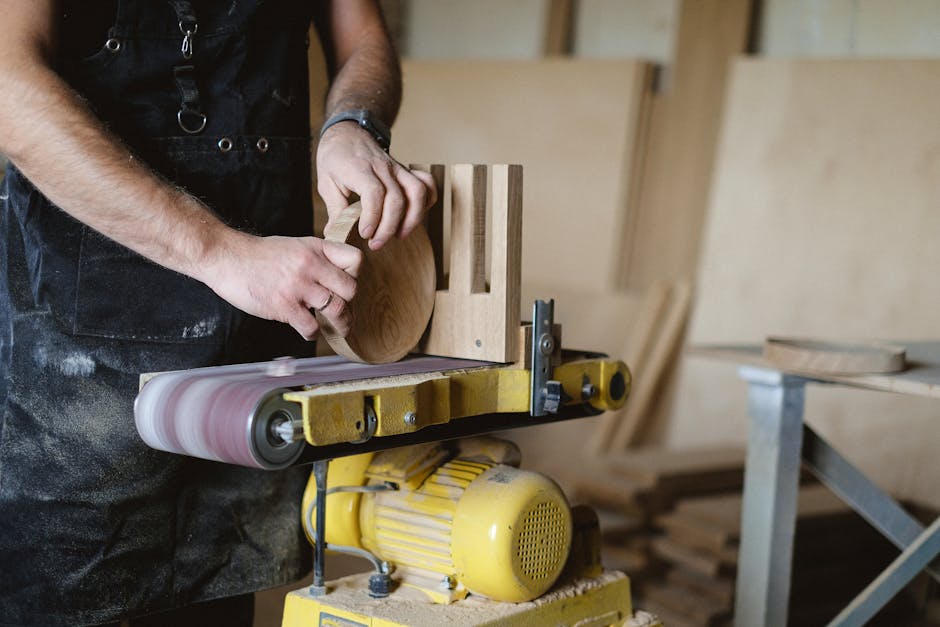 Anonymous male master using belt and disc sander to shape piece of wood while working in professional carpentry with stack of wood